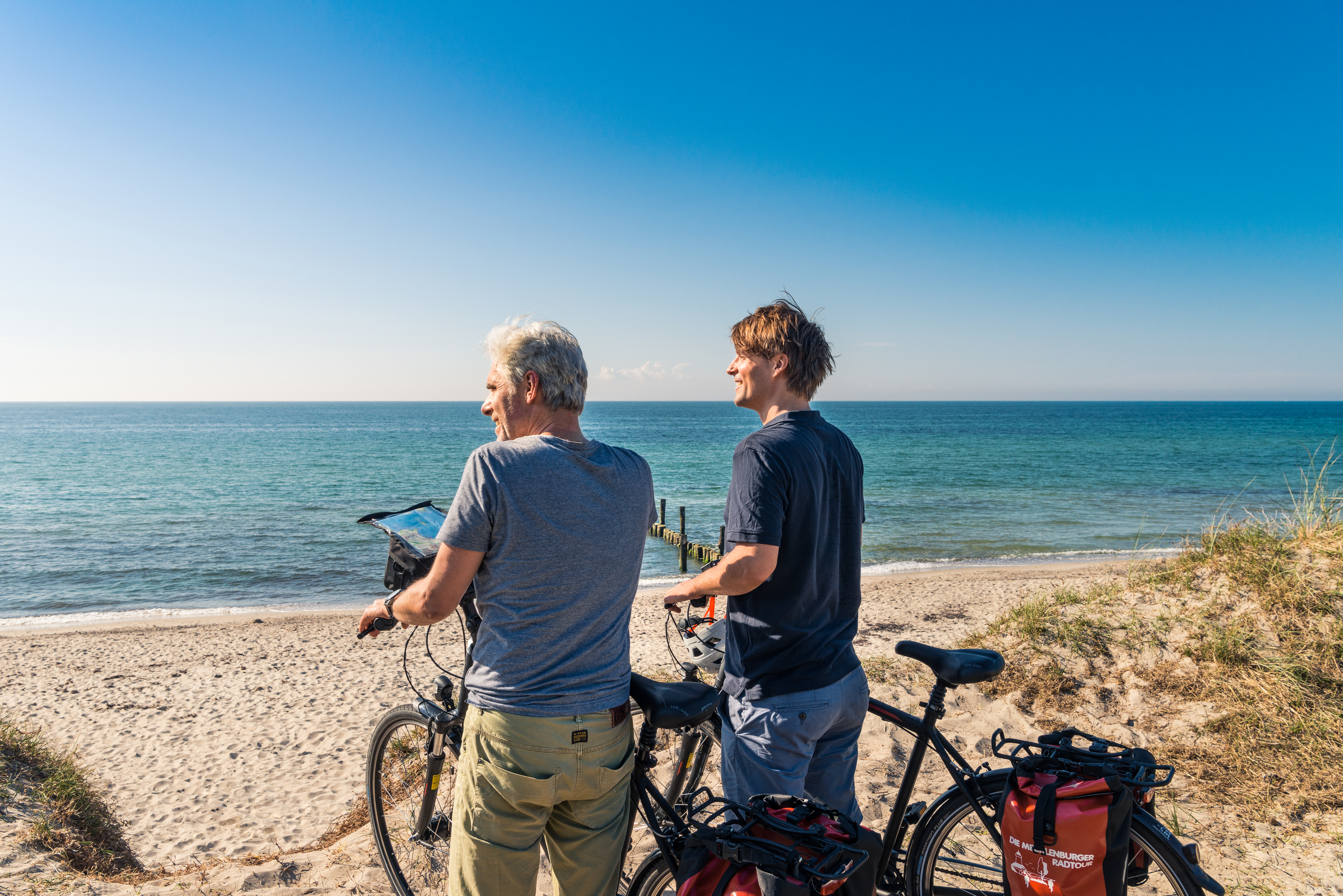 Zwei Männer stehen mit dem Rücken zur Kamera und zum Meer gerichtet am Strand mit ihren Fahrrädern und blicken auf das türkise Wasser. Sie sehen zufrieden aus